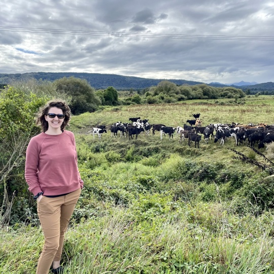 Kaitlyn smiling at the camera while standing in a field with several cows in the distance behind her.